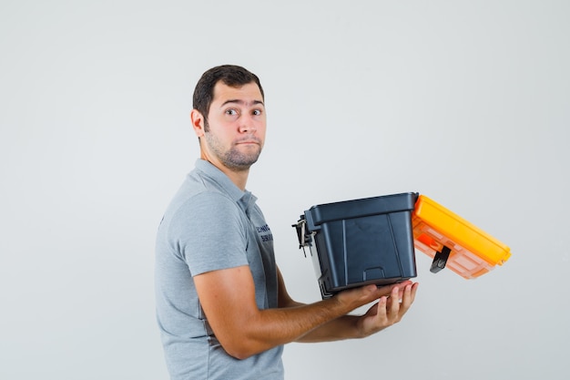 Jeune technicien en uniforme gris tenant une boîte à outils ouverte et à la recherche de sérieux.