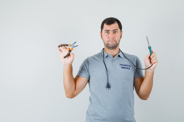 Jeune technicien en uniforme gris tenant une perceuse dans une main, une pince dans une autre main et à la recherche de sérieux.