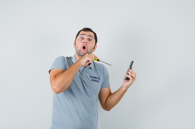 Jeune technicien en uniforme gris tenant un tournevis et essayant d'ouvrir le dos de son téléphone et à la stupéfaction