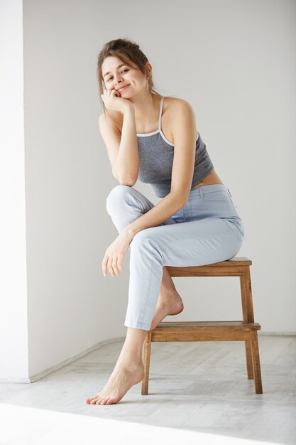 Jeune tendre belle femme souriante assise sur une chaise sur un mur blanc.