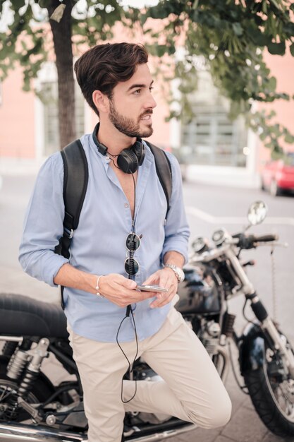 Jeune touriste mâle avec smartphone debout devant la moto