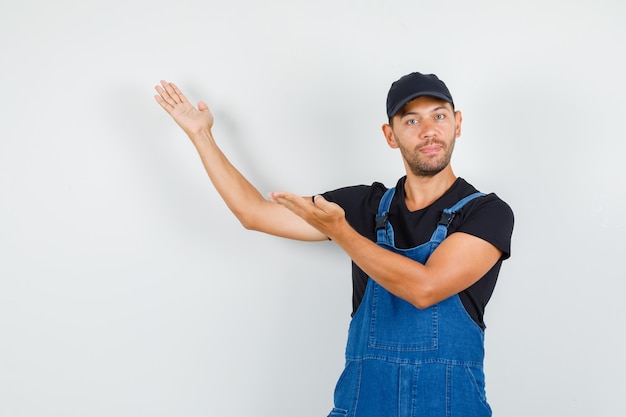 Photo gratuite jeune travailleur accueillant ou montrant quelque chose avec les paumes ouvertes en uniforme, vue de face.