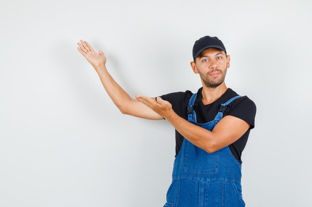 Jeune travailleur accueillant ou montrant quelque chose avec les paumes ouvertes en uniforme, vue de face.