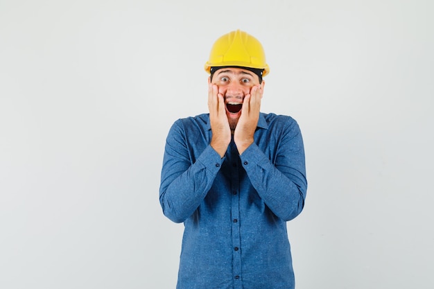 Jeune travailleur en chemise, casque tenant les mains sur les joues et à la surprise