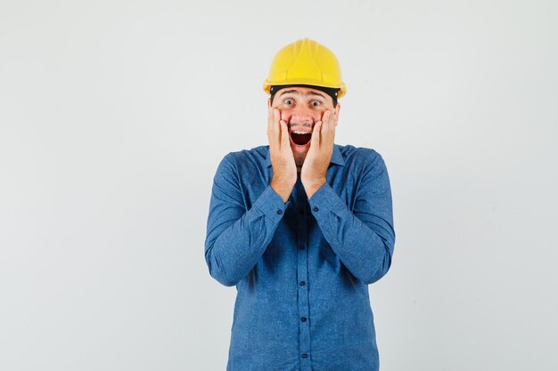 Jeune travailleur en chemise, casque tenant les mains sur les joues et à la surprise