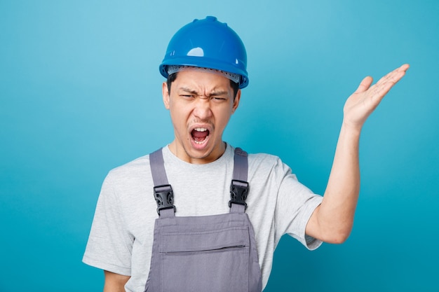 Jeune travailleur de la construction en colère portant un casque de sécurité et l'uniforme montrant des cris de main vide