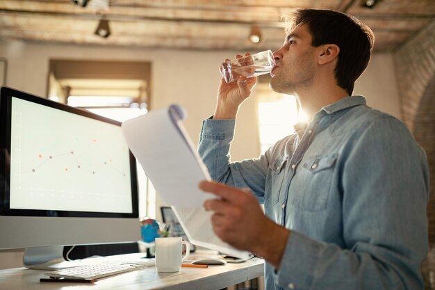 Jeune travailleur indépendant buvant un verre d'eau tout en travaillant sur de la paperasse au bureau