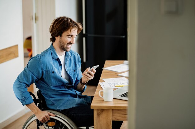 Jeune travailleur indépendant heureux dans la messagerie texte en fauteuil roulant sur téléphone mobile tout en travaillant à la maison