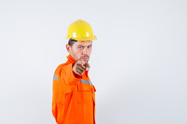 Jeune travailleur pointant vers l'avant en uniforme, casque et à la recherche de sérieux.