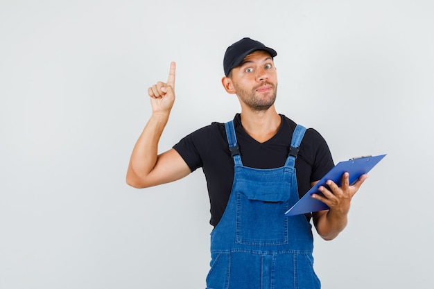 Jeune travailleur pointant vers le haut et tenant le presse-papiers en uniforme, vue de face.