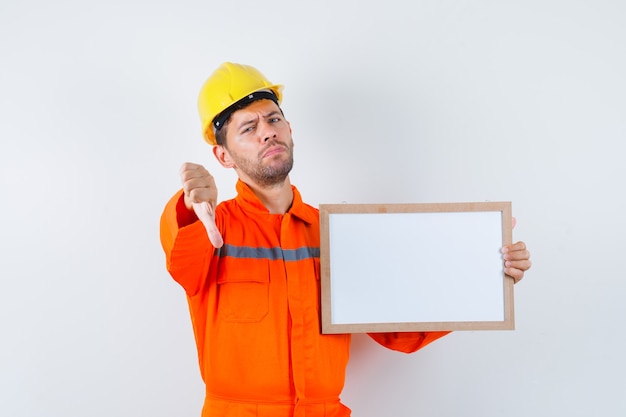 Jeune travailleur tenant un cadre vierge, montrant le pouce vers le bas en uniforme, casque.