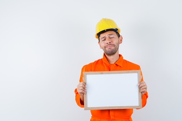 Jeune travailleur tenant un cadre vierge en uniforme, casque et air triste.