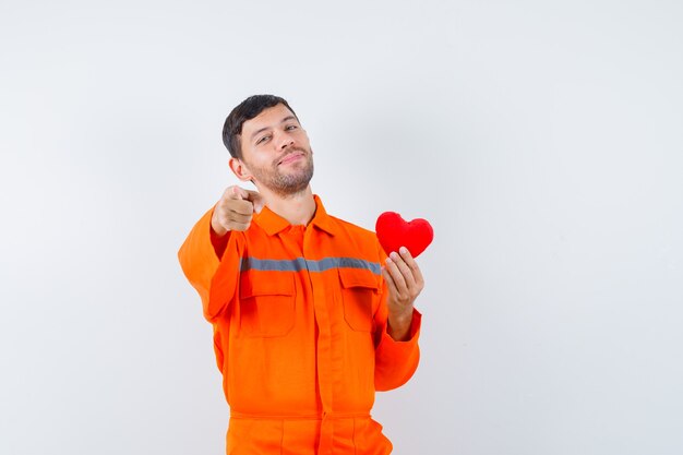 Jeune travailleur tenant un coeur rouge, pointant vers l'avant en uniforme et à la bonne humeur.