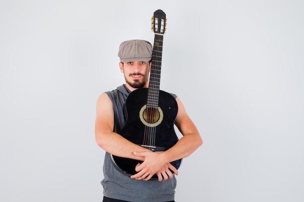 Jeune travailleur tenant une guitare avec les deux mains en t-shirt et casquette gris et ayant l'air heureux