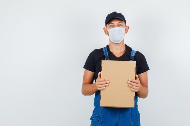 Jeune travailleur en uniforme, masque tenant une boîte en carton et regardant grave, vue de face.