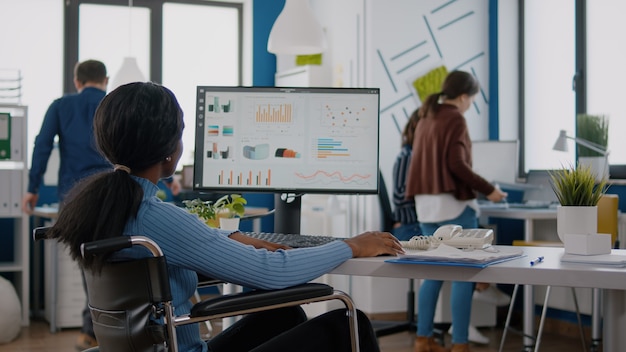 Jeune travailleuse handicapée analysant les informations statistiques assise dans un fauteuil roulant devant un ordinateur tapant un projet financier sur le lieu de travail de démarrage