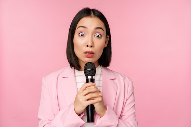 Jeune vendeuse asiatique dame de bureau avec costume tenant un microphone et regardant choqué à la caméra parlant donnant un discours debout sur fond rose Espace de copie