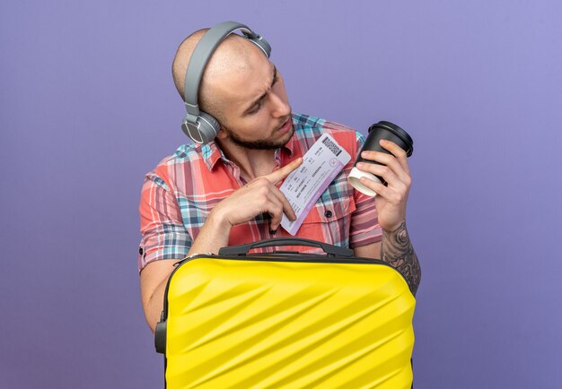 Jeune voyageur désemparé sur un casque tenant un billet d'avion et regardant une tasse en papier debout derrière une valise isolée sur un mur violet avec espace pour copie