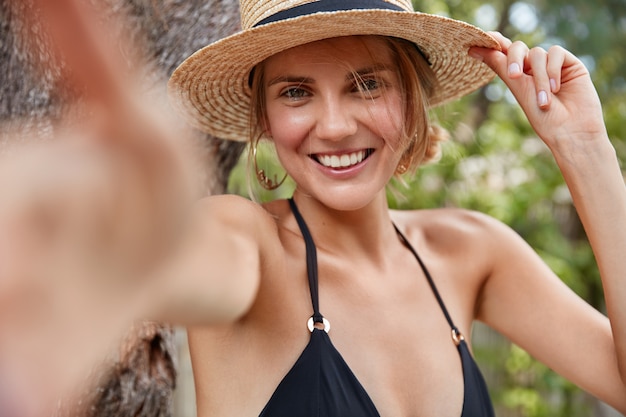 Jeune voyageur féminin souriant attrayant en chapeau de paille et bikini, fait selfie sur fond tropical, satisfait de passer des vacances d'été à l'étranger dans un pays exotique. Concept de beauté et de repos