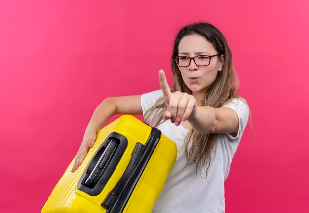 Jeune voyageur femme en t-shirt blanc tenant une valise de voyage montrant l'index d'avertissement avec un visage sérieux debout sur un mur rose