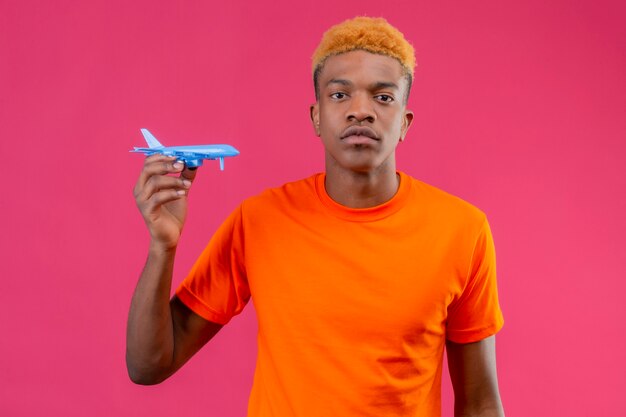 Jeune voyageur garçon portant un t-shirt orange tenant un avion jouet avec une expression de confiance grave debout sur un mur rose