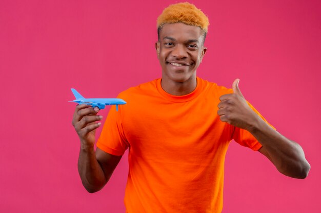 Jeune voyageur garçon portant un t-shirt orange tenant un avion jouet souriant montrant les pouces vers le haut debout sur un mur rose