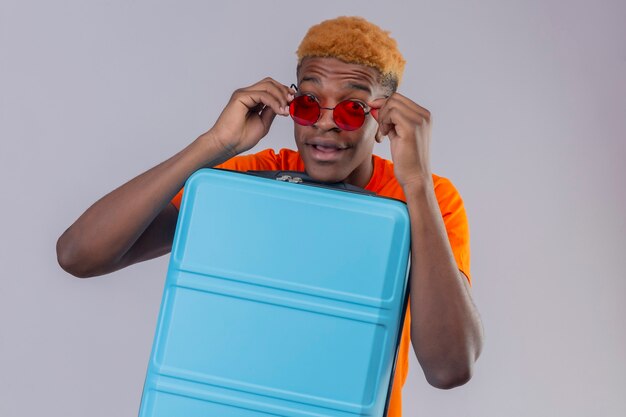 Jeune voyageur garçon portant un t-shirt orange tenant une valise intriguée en prenant ses lunettes de soleil debout sur un mur blanc