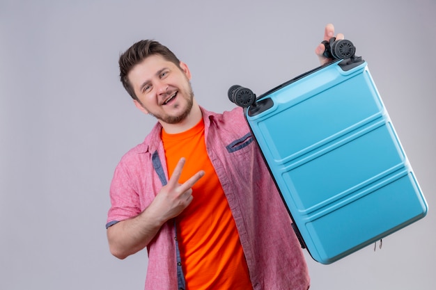 Photo gratuite jeune voyageur homme tenant une valise bleue regardant la caméra en souriant joyeusement positif et heureux montrant le numéro deux ou signe de la victoire debout sur fond blanc