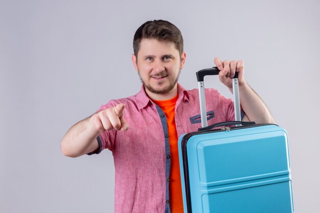 Jeune voyageur homme tenant valise regardant la caméra confiant pointant avec le doigt debout sur fond blanc