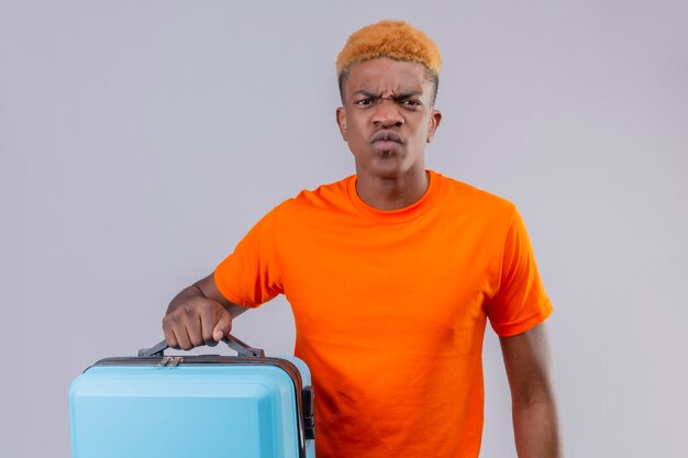 Jeune voyageur mécontent garçon portant un t-shirt orange tenant une valise avec un visage fronçant debout sur un mur blanc
