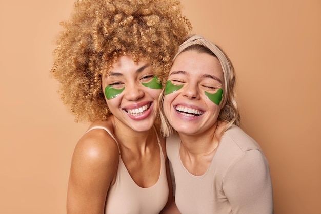 De jeunes amies positives sourient largement s'amusent ensemble appliquent des patchs d'hydrogel vert sous les yeux subissent des procédures de beauté habillées avec désinvolture posent sur fond beige Concept de soins de la peau