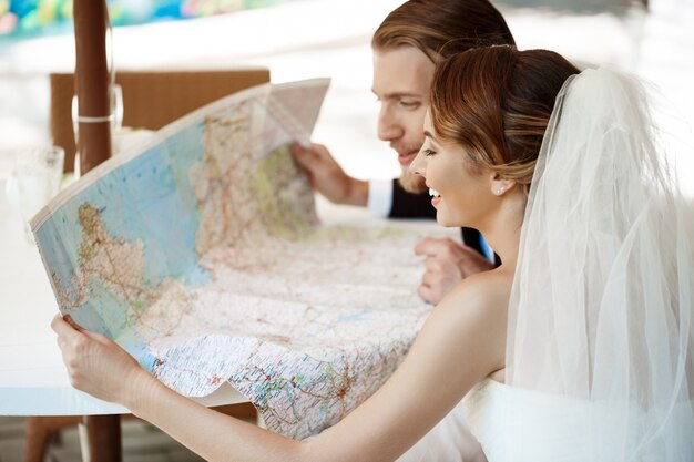 Jeunes beaux jeunes mariés souriant, choisissant un voyage de noces, en regardant la carte.