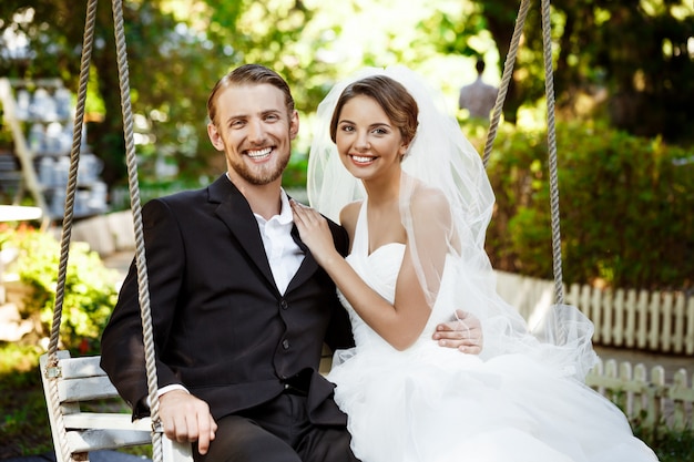 Photo gratuite jeunes beaux jeunes mariés souriant, riant, assis sur une balançoire dans le parc.