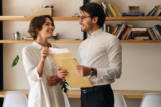 Jeunes collègues de travail attrayants debout avec une lettre d'enveloppe tout en se regardant joyeusement dans un bureau moderne