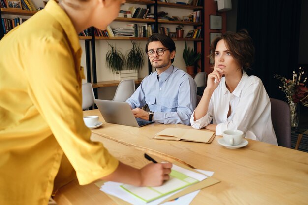 Jeunes collègues de travail réfléchis assis au bureau avec un ordinateur portable tout en écoutant attentivement le patron au travail dans un bureau moderne