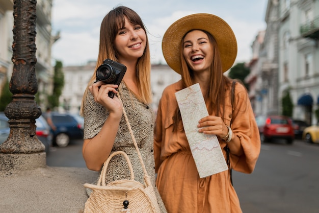 Jeunes femmes élégantes voyageant ensemble vêtues de robes et d'accessoires à la mode du printemps s'amusant à prendre des photos sur une caméra tenant une carte