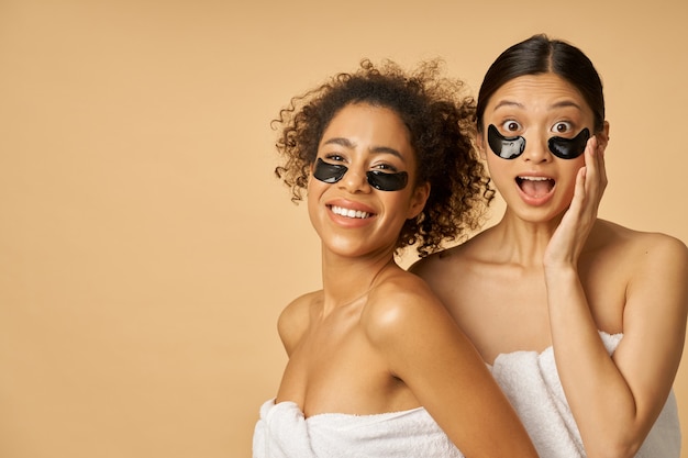 Jeunes femmes enveloppées dans des serviettes posant avec du gel hydro noir appliqué sous les cache-œil sur la peau du visage