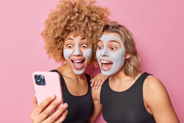 Des jeunes femmes heureuses et surprises regardent l'écran du smartphone pour que le selfie garde la bouche ouverte, applique un masque facial de boue, debout l'une à côté de l'autre, vêtue de t-shirts noirs décontractés isolés sur fond rose