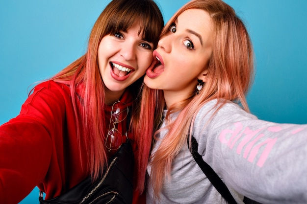 Jeunes femmes hipster folles faisant selfie au mur bleu, émotions drôles surpris, longs cheveux roses, tenues décontractées.