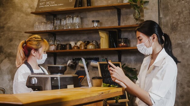 Les jeunes femmes portent un masque facial en libre-service utilisent un téléphone portable sans contact au restaurant.