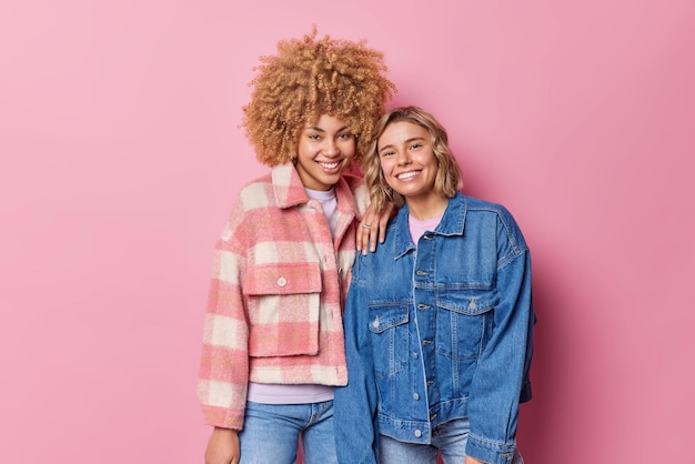 Des jeunes femmes satisfaites sourient agréablement debout l'une à côté de l'autre vêtues de vestes à la mode étant de bonne humeur isolées sur fond rose Deux amies joyeuses se rencontrent pour poser pour la photo