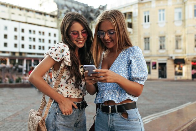 Jeunes filles bronzées en pantalons en denim et chemisiers élégants regardent l'écran du téléphone violet Des femmes blondes et brunes en lunettes de soleil colorées sourient et parlent à l'extérieur