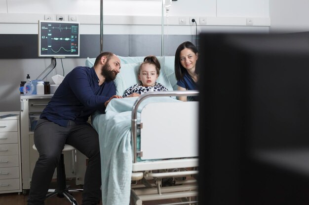 Jeunes parents et fille malade regardant la chaîne de télévision assis dans la salle de consultation de la clinique pédiatrique. La famille passe du temps ensemble dans la chambre du patient de la clinique tout en profitant d'une émission de télévision.