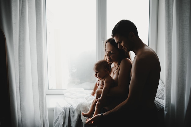 De jeunes parents tiennent un nouveau-né devant une fenêtre lumineuse dans une pièce sombre