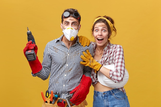 Les jeunes travailleurs industriels à la recherche avec les yeux buggés en se tenant debout contre un mur blanc jaune. Beau couvreur professionnel en masque de protection tenant une perceuse électrique ayant un kit d'instruments