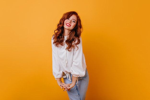 Jocund caucasin lady smiling sur mur orange. Photo intérieure d'une femme frisée raffinée dans une élégante blouse blanche.