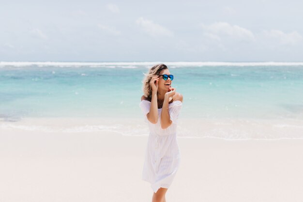 Jocund femme en tenue blanche romantique debout sur la mer. Rire femme extatique à lunettes de soleil, passer la journée d'été à l'océan.