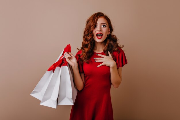 Jocund jeune femme aux cheveux longs posant après le shopping. Dame de gingembre insouciante tenant des sacs de magasin.