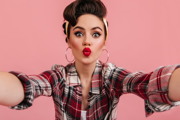 Photo gratuite jocund pin-up posant en chemise à carreaux rouge. femme étonnée prenant selfie avec l'expression du visage embrassant.