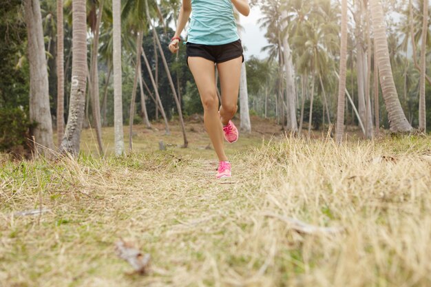 Jogger femme caucasienne avec beau corps en forme en cours d'exécution sur l'herbe dans la forêt tropicale. Jeune coureuse portant un haut de sport bleu et un short noir exerçant à l'extérieur par une journée ensoleillée.